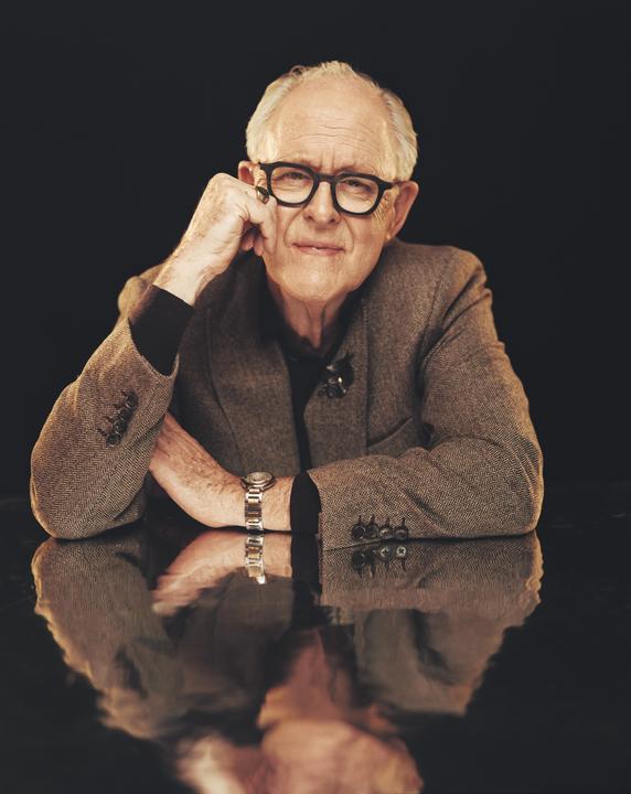 A man wearing glasses and a tweed jacket sits at a reflective table, resting his chin on his hand.