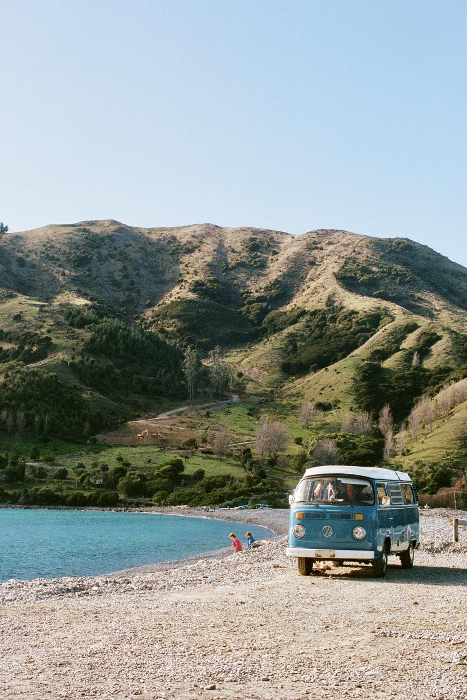 A blue van is parked on a gravel beach next to a body of water with green hills in the background.