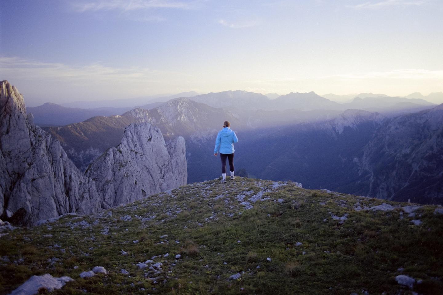 A woman stands on a grassy mountain peak, looking out at a vast mountain range under a hazy sky.