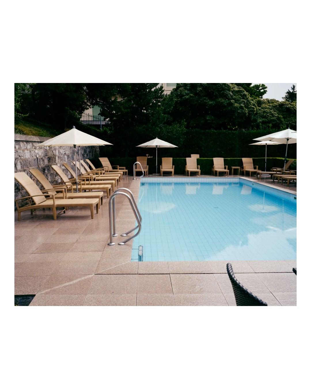 A swimming pool with lounge chairs and umbrellas is surrounded by trees and a stone wall.