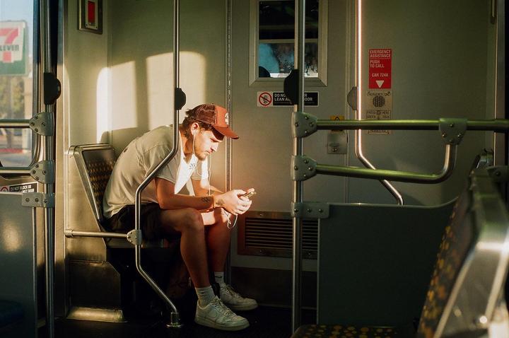 A man wearing a baseball cap and earphones sits on a train, looking at his phone.