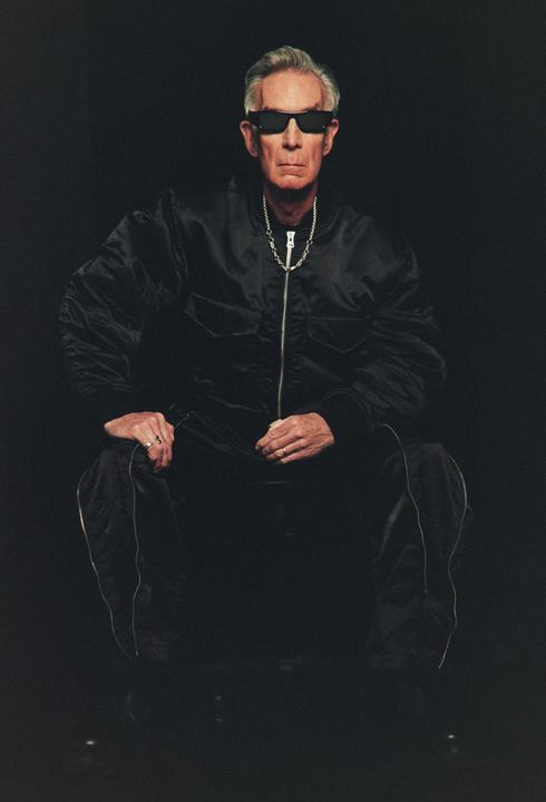 A man wearing sunglasses and a black jacket is crouching in a dark setting.