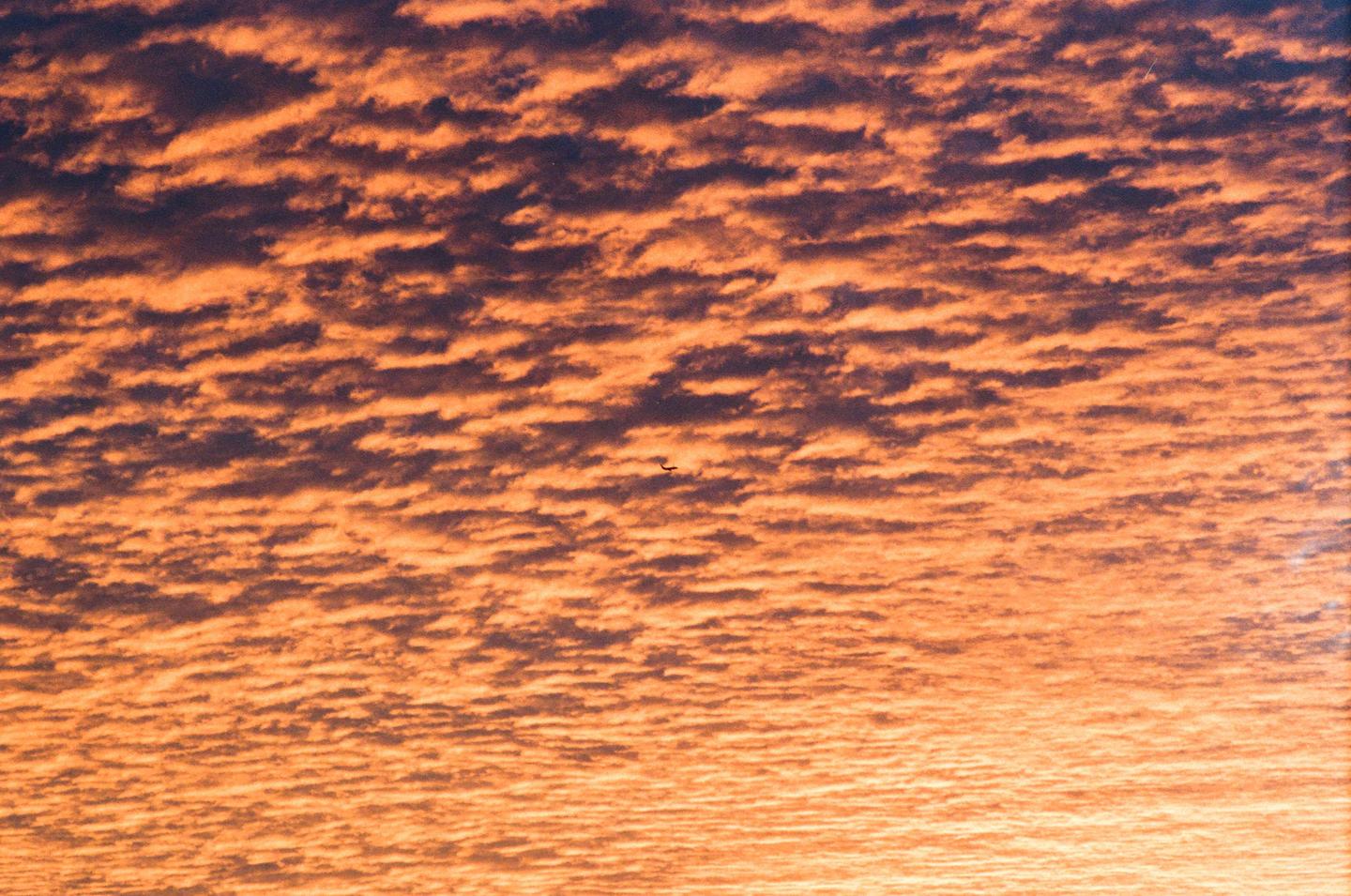 A small airplane flies through a sky filled with orange and dark clouds during sunset.
