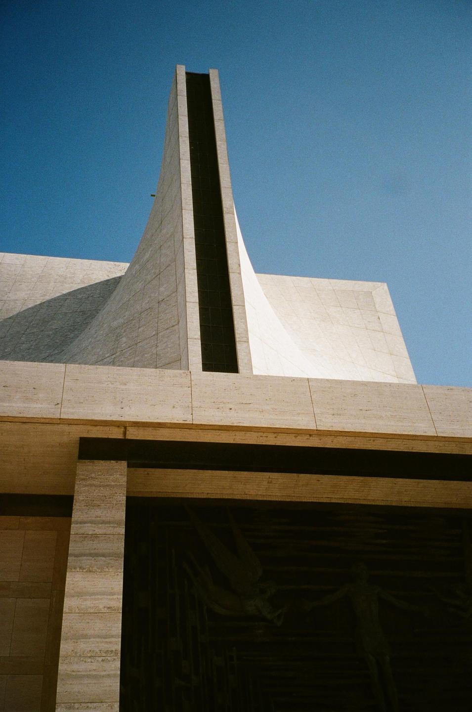 A low-angle view shows the concrete facade of a modern building against a clear blue sky.