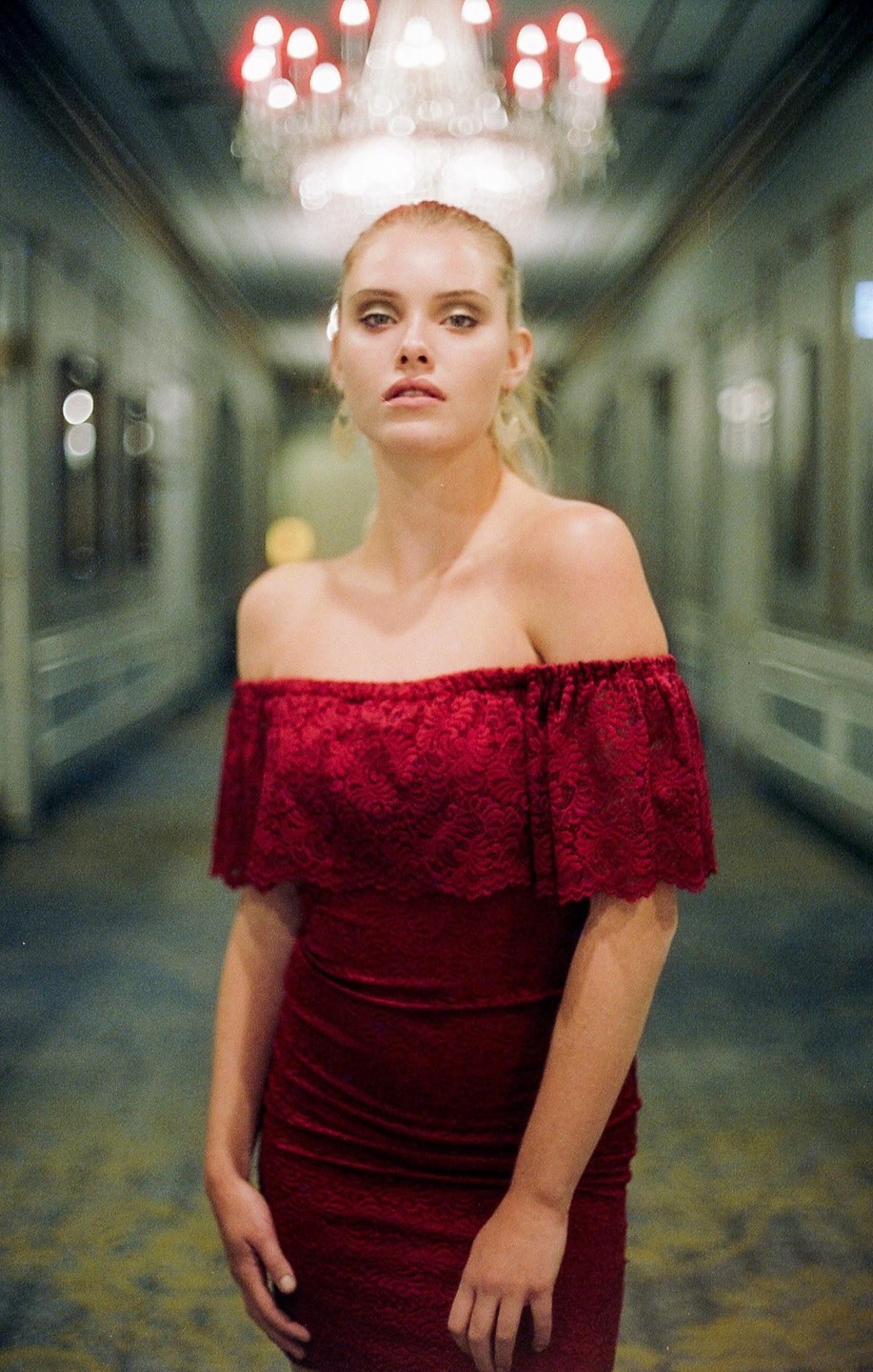 A woman in a red dress stands in a hallway with a chandelier overhead.