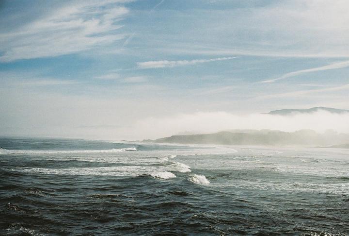 Waves crash on a beach with a hazy coastline and mountains in the background under a blue sky.