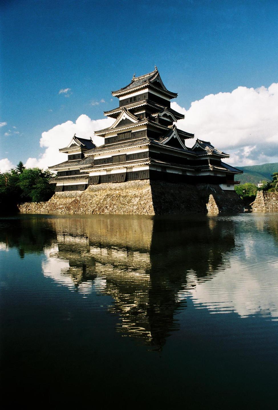 A traditional Japanese castle with multiple dark tiers and white trim is reflected in the still water of a moat under a blue sky with white clouds.