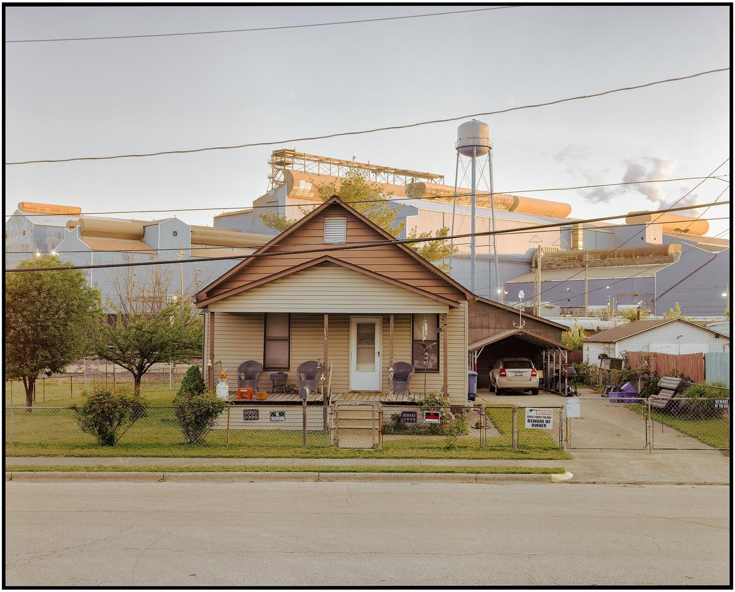 A small house with a porch and a carport sits in front of a large industrial building with a water tower.
