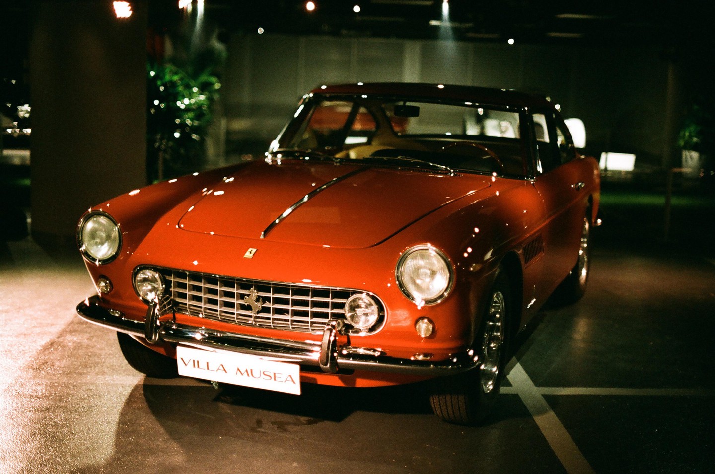 A red vintage car with a "VILLA MUSEA" license plate is parked indoors at night.