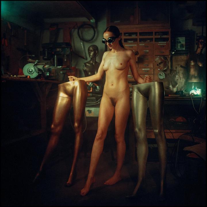 A nude woman wearing goggles stands between two mannequin legs in a workshop.
