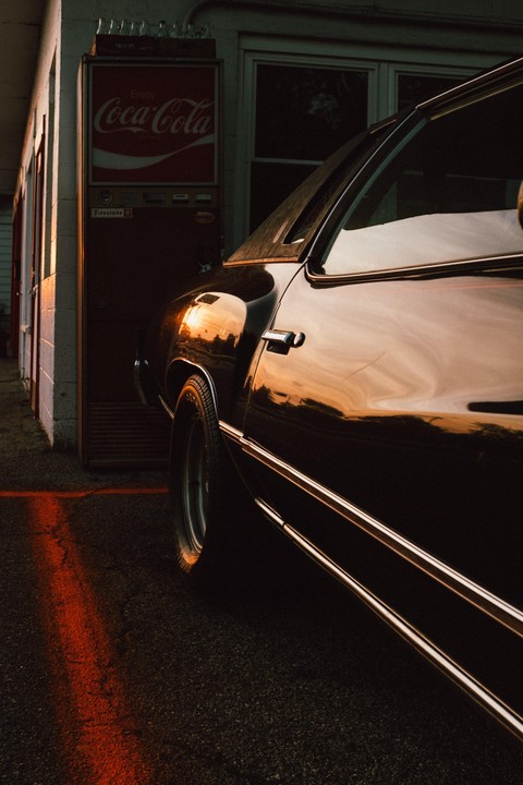 A dark car is parked next to a Coca-Cola vending machine with reflections of the sunset on its body.