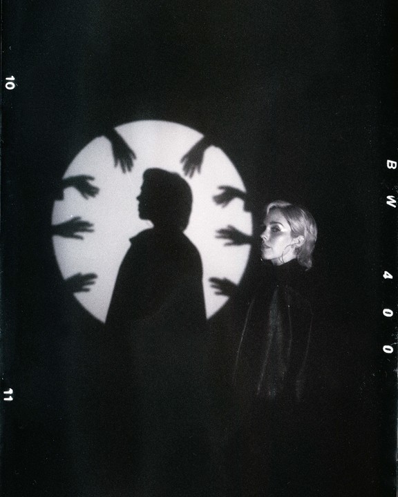 A woman stands next to a large circular light with a silhouette of a person and multiple hands projected onto it.
