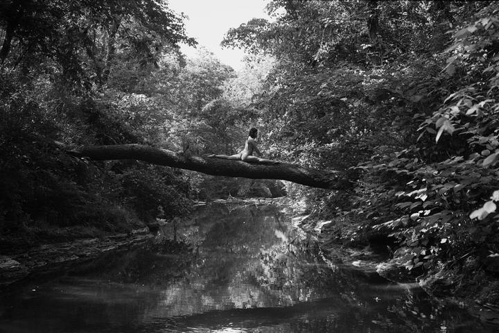A woman sits on a fallen tree trunk that spans a narrow river in a forest.