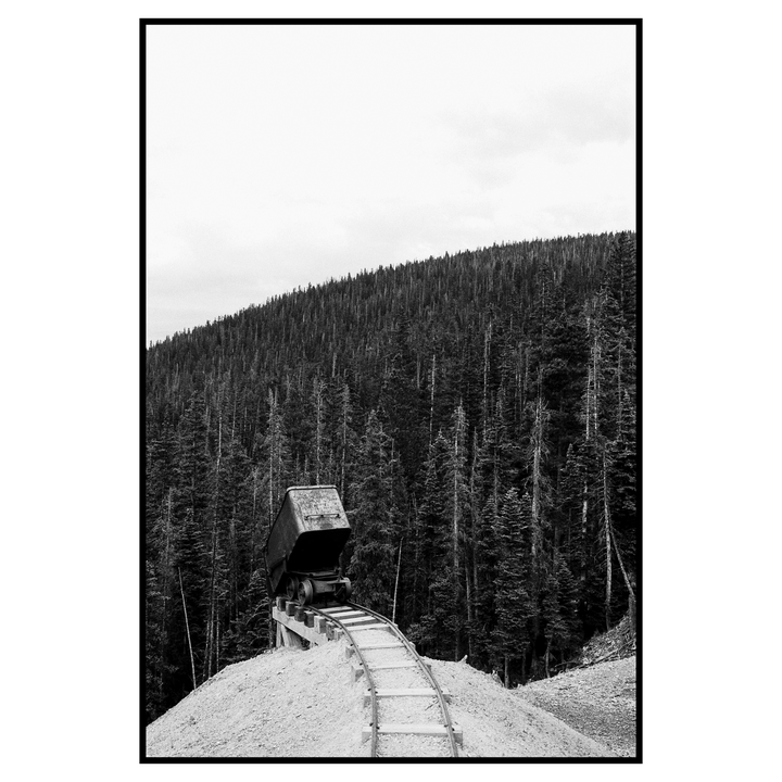 A mining cart sits on a track on a hill of dirt with a dense forest in the background.