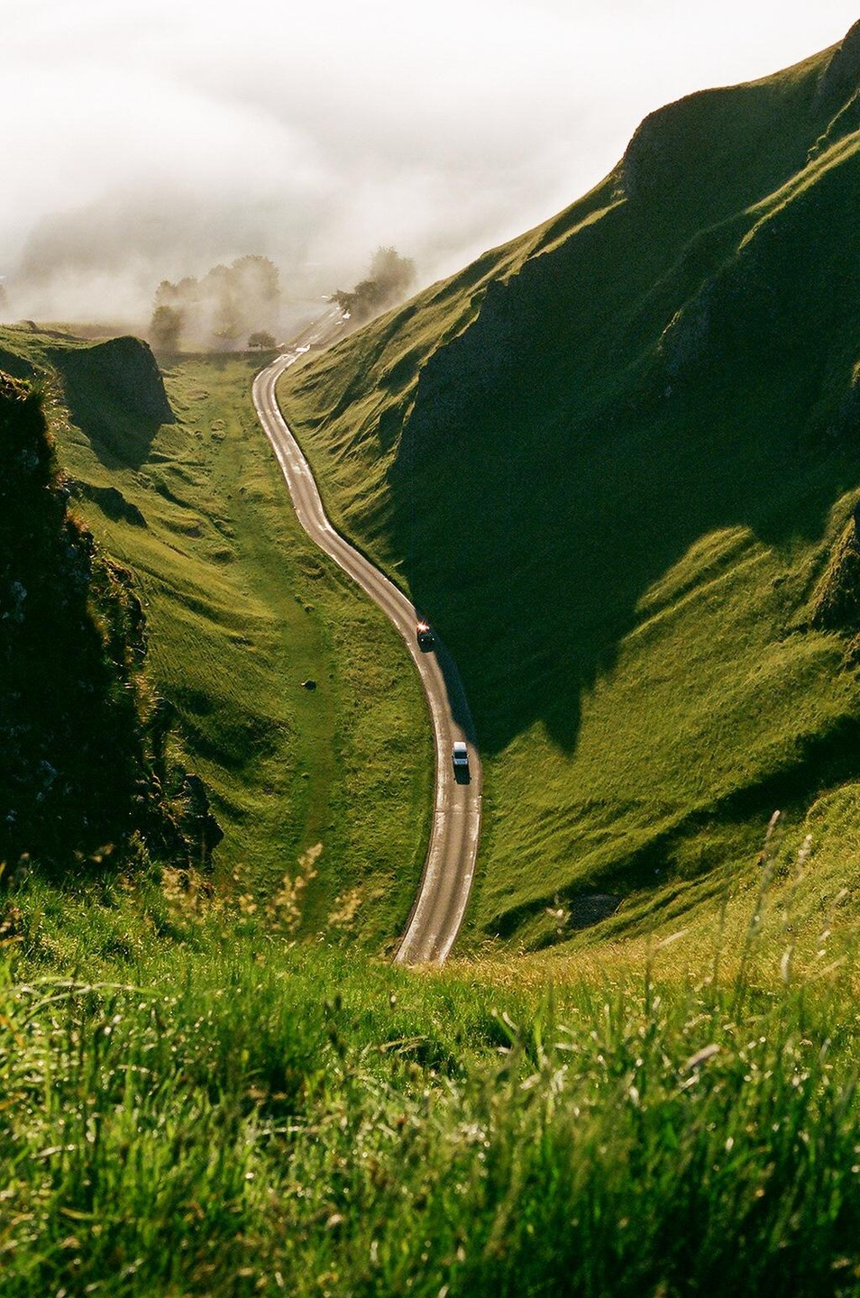 A winding road with two cars travels through a green valley with fog in the distance.