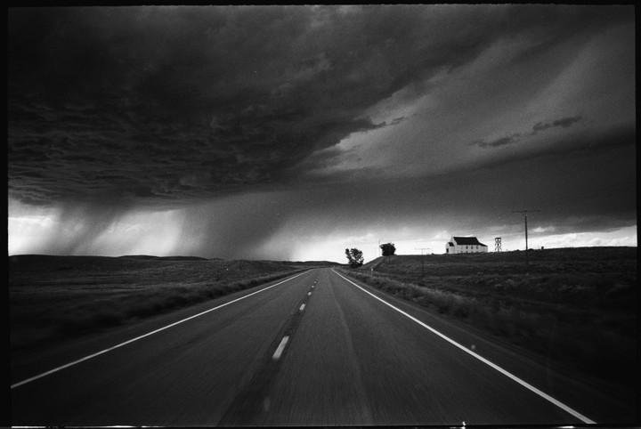 A long road leads toward a white building on a hill under a dark, stormy sky with rain falling in the distance.