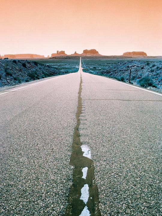 A long, straight road leads to distant rock formations under an orange sky.