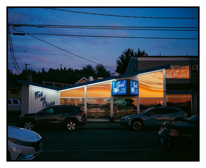 A fish and chips restaurant with large windows reflects the sunset sky, with cars parked on the street in front.