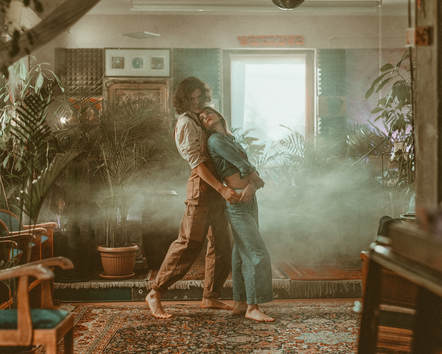A couple embraces in a hazy room filled with plants and patterned rugs.
