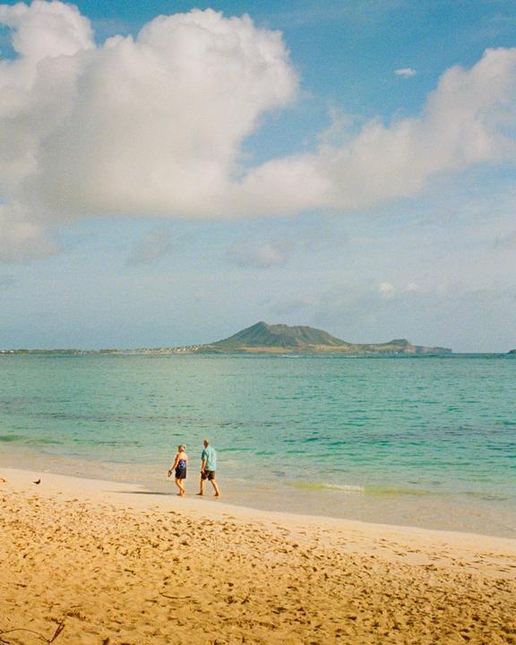 A couple walks along a sandy beach with a mountain in the distance under a cloudy sky.
