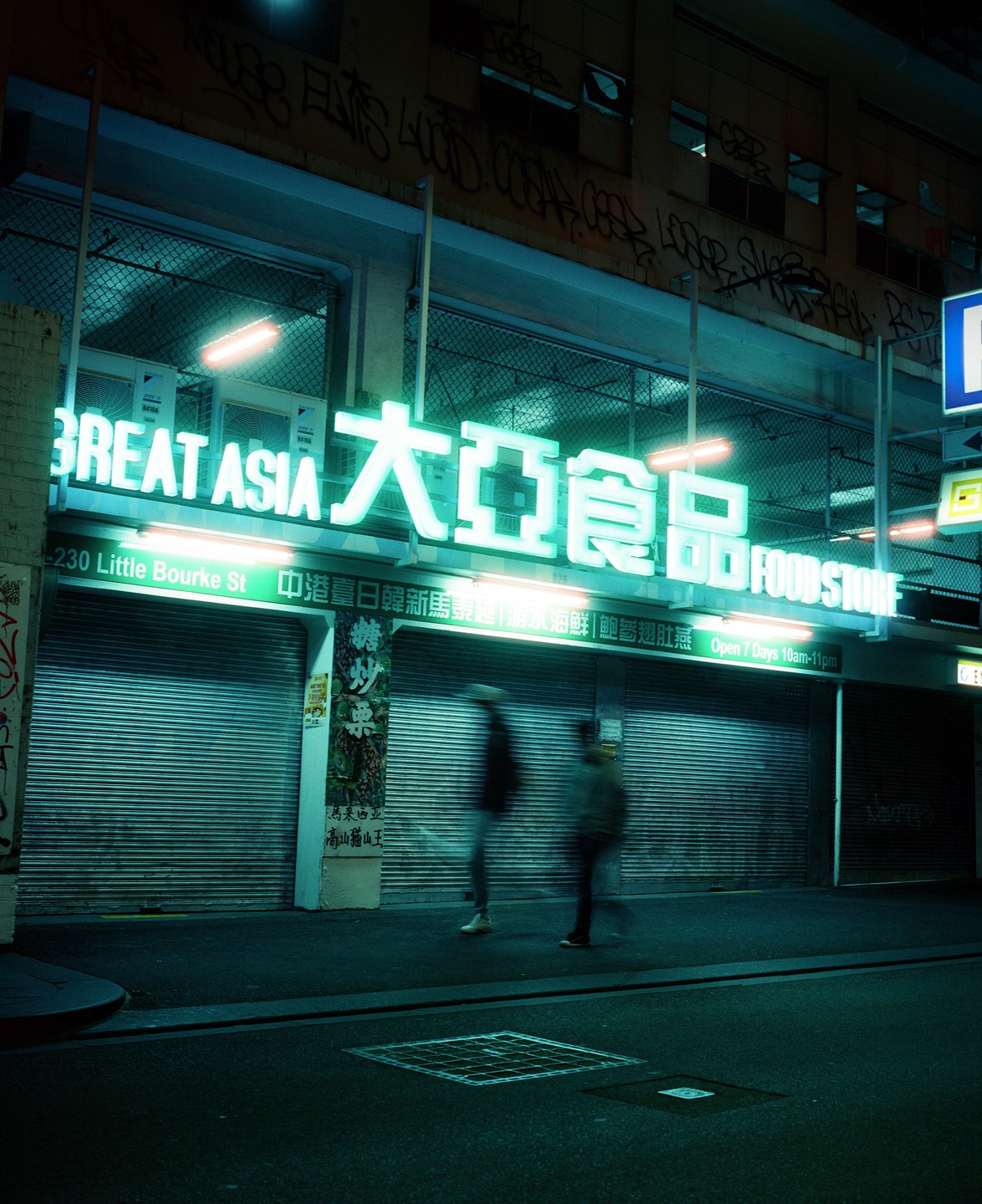 Two blurry people walk past a closed Great Asia Food Store with neon signs at night.