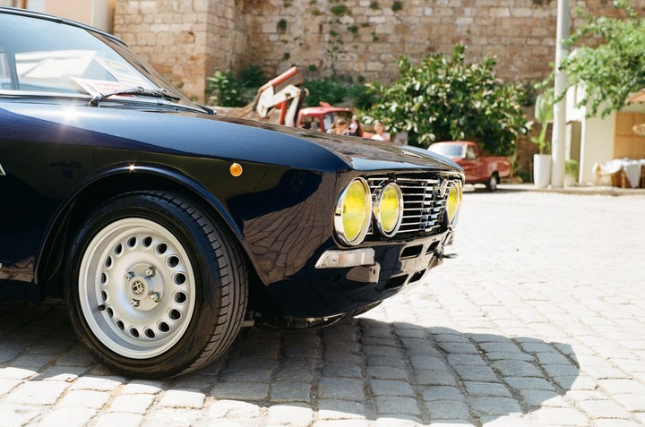 A dark blue vintage car with yellow headlights is parked on a cobblestone street.