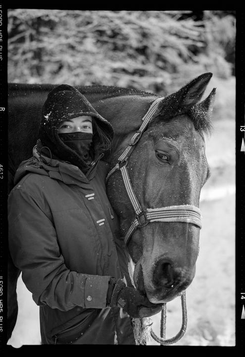 A woman wearing a hooded jacket and face covering stands next to a horse in a snowy setting.