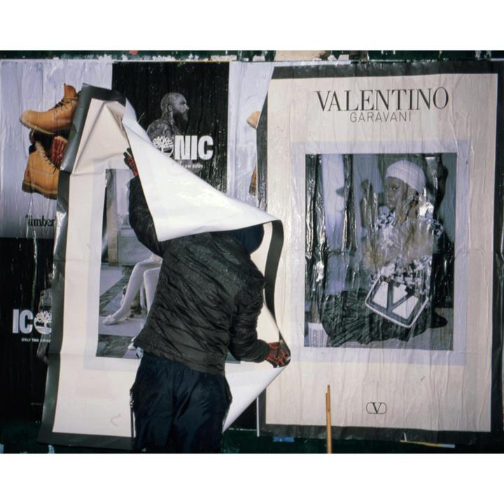 A person is peeling a large poster off a wall covered with other posters.