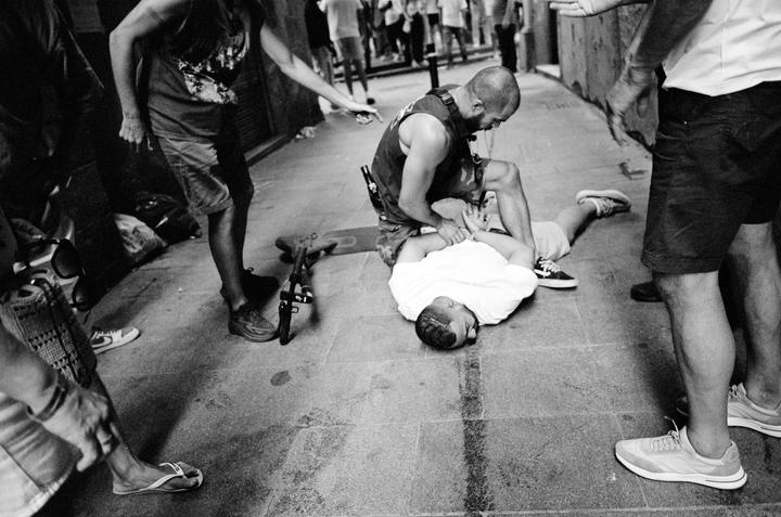 A man is on the ground with another man kneeling over him while other people stand around them on a street.