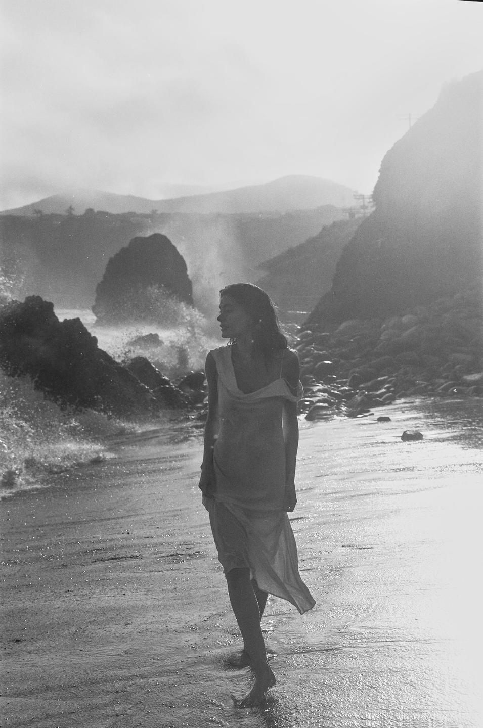 A woman in a dress stands on a rocky beach with waves crashing behind her.