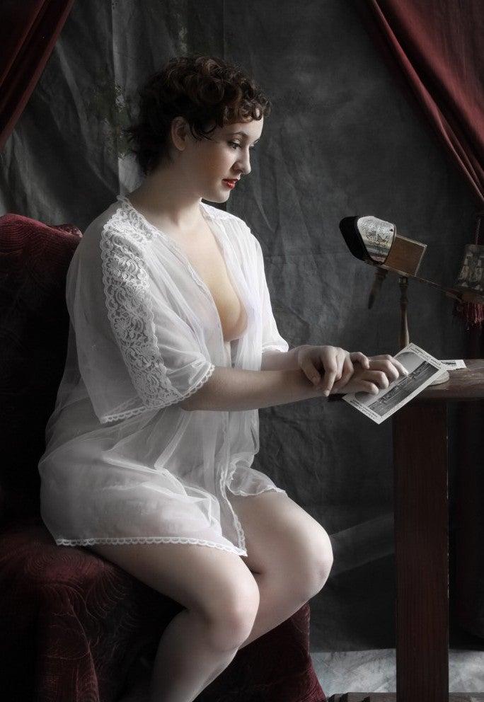 A woman in a sheer white robe sits on a red velvet chair, looking at a photograph.