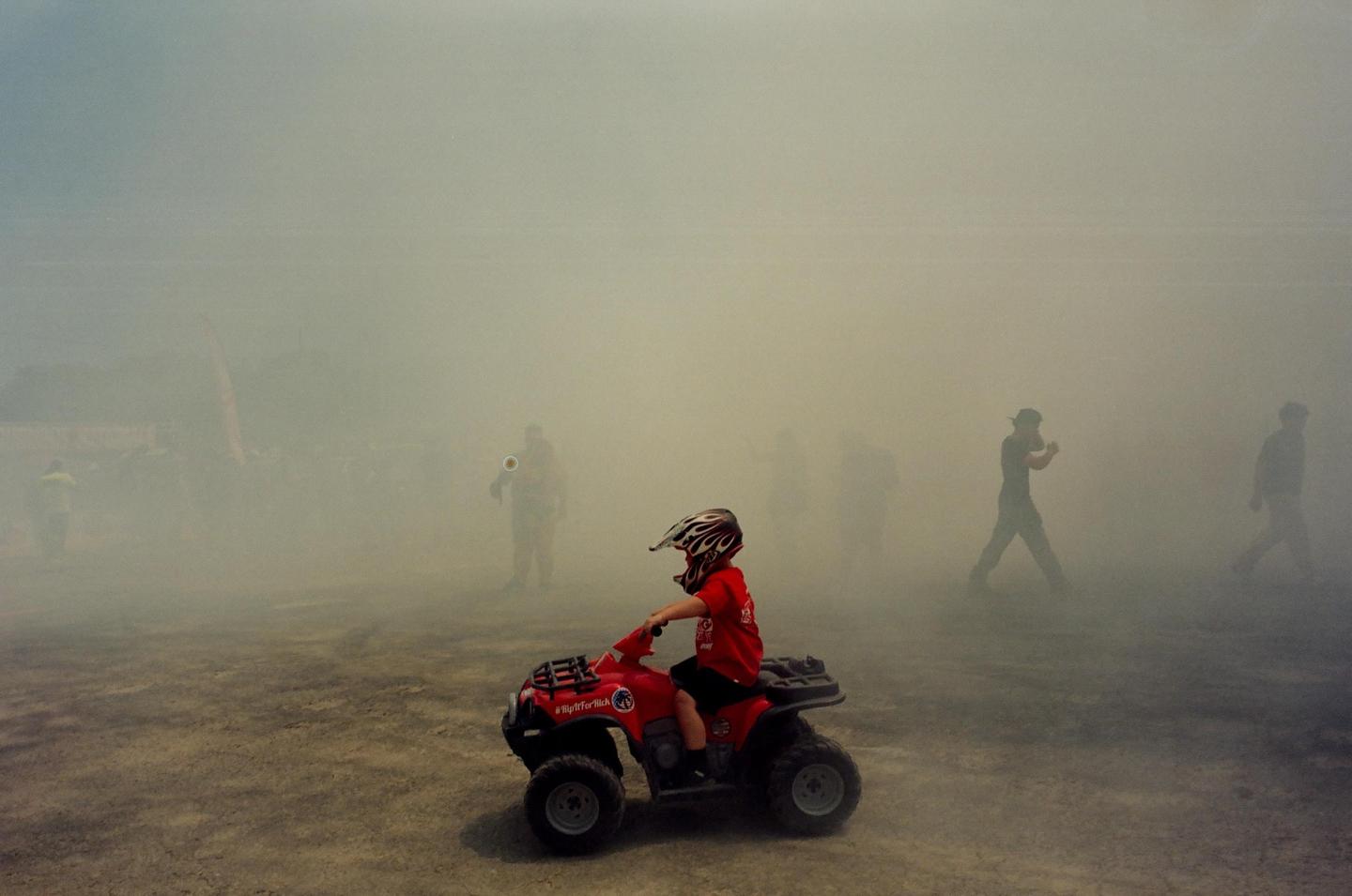 A child in a helmet and red shirt sits on a red toy ATV in a hazy, dusty area with several blurred figures in the background.