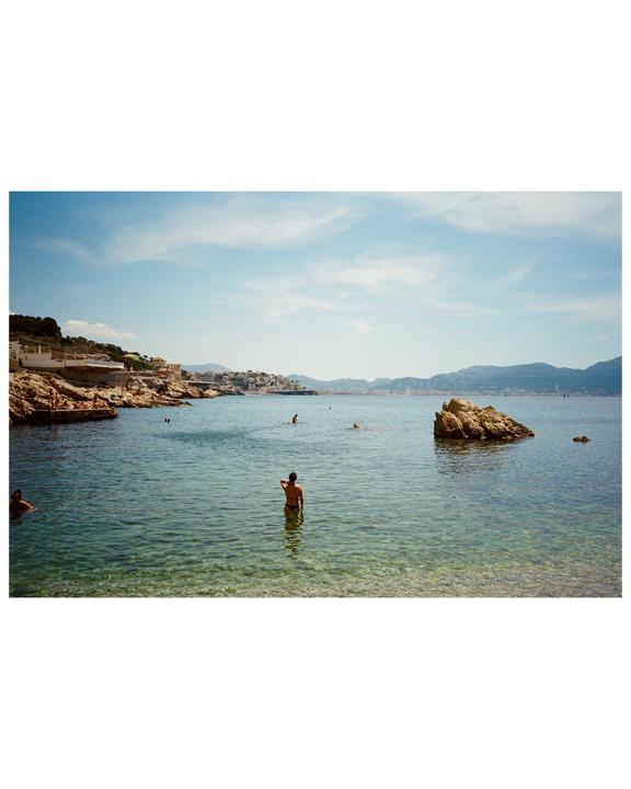 A person stands in clear water with a rocky coastline and distant city under a blue sky.