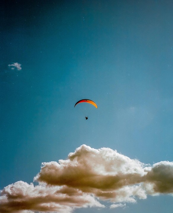 A paraglider with an orange and red parachute flies above a large cloud in a blue sky.