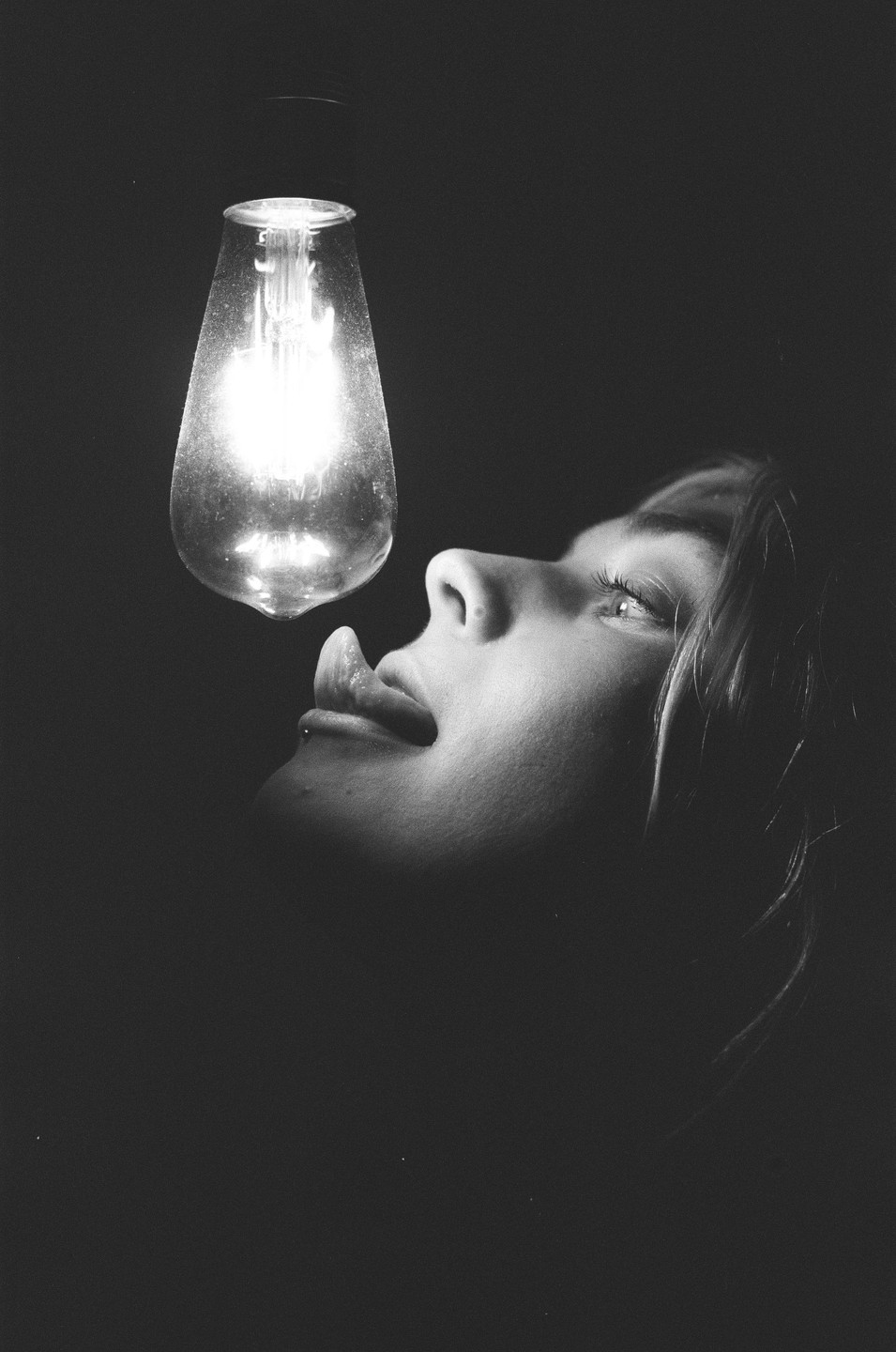 A woman with her tongue out looks up at a glowing lightbulb in a dark setting.