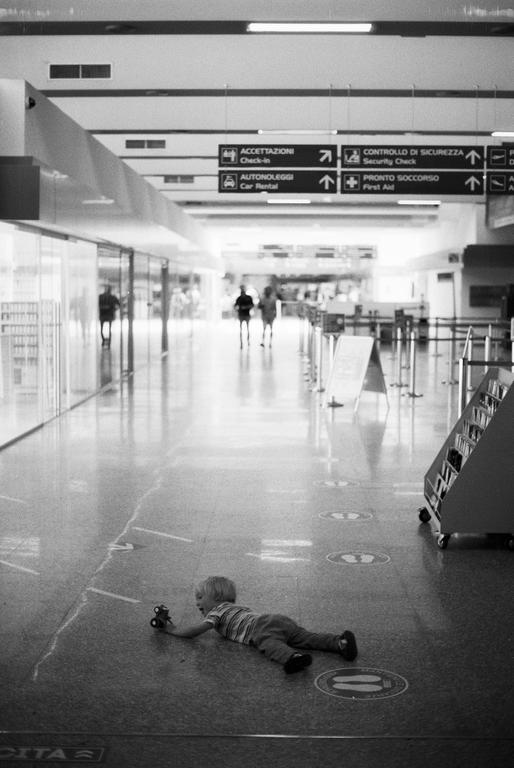 A child lies on the floor of an airport terminal, playing with a toy car, while two adults walk in the background.