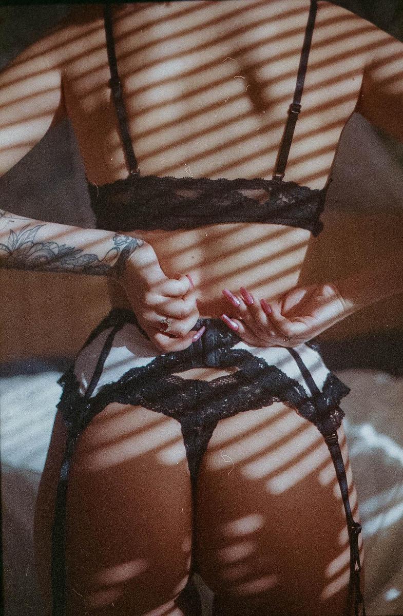A woman in lingerie is seen from behind, fastening her garter belt, with shadows from blinds cast across her body.