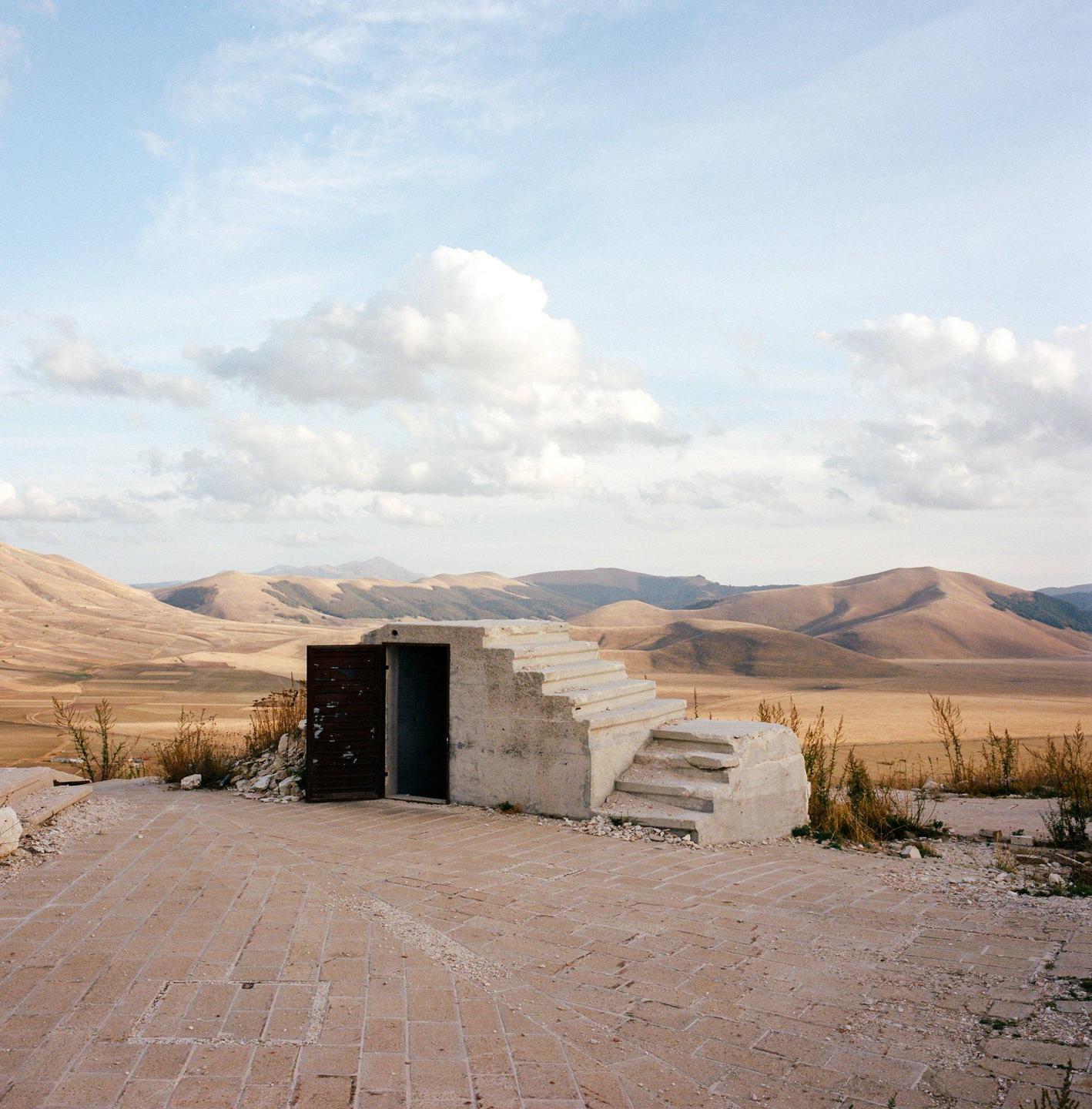 A small concrete structure with an open door and a set of stairs sits on a paved area overlooking a vast, hilly landscape under a cloudy sky.
