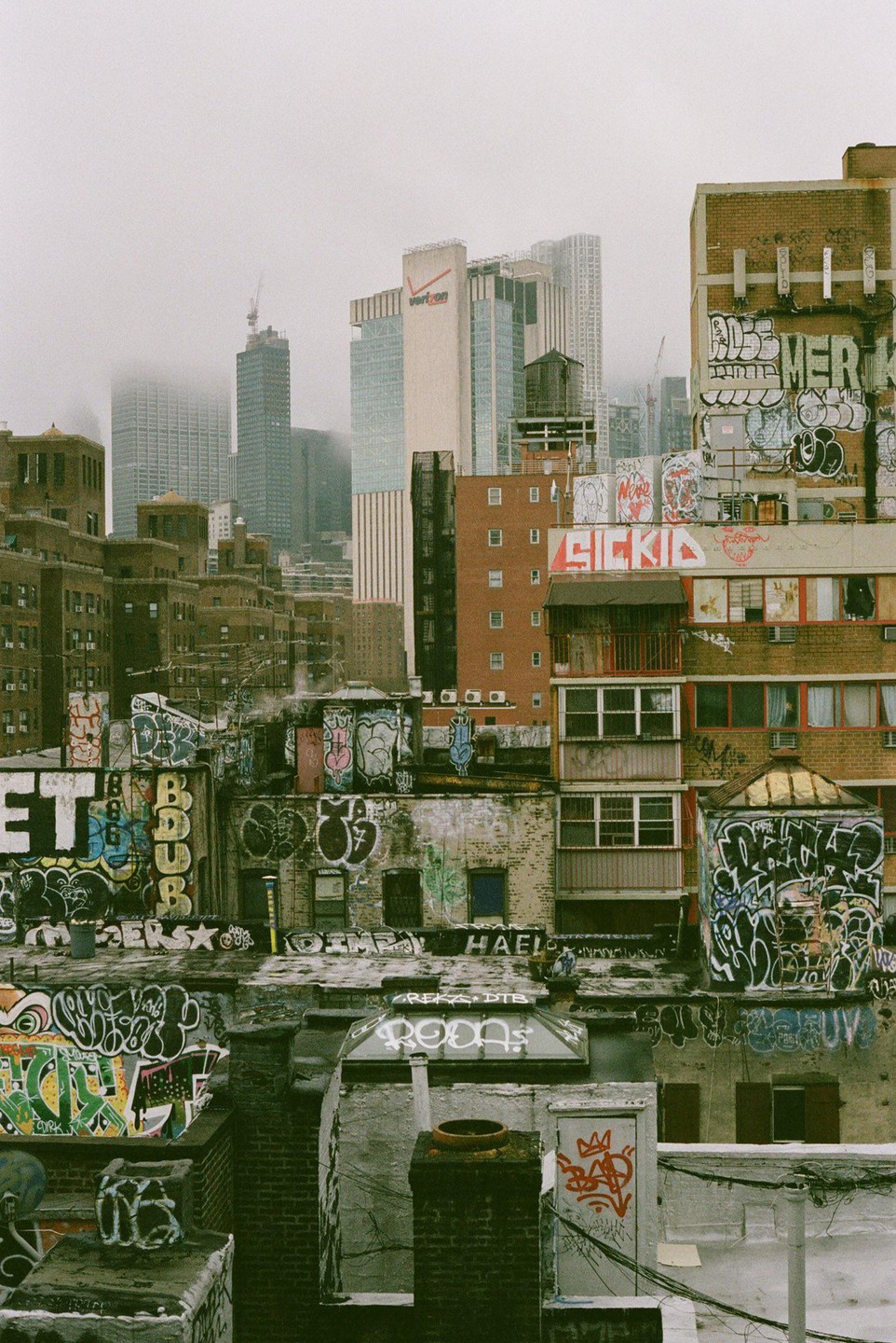 A dense urban landscape features buildings covered in graffiti, with taller structures and a hazy sky in the background.