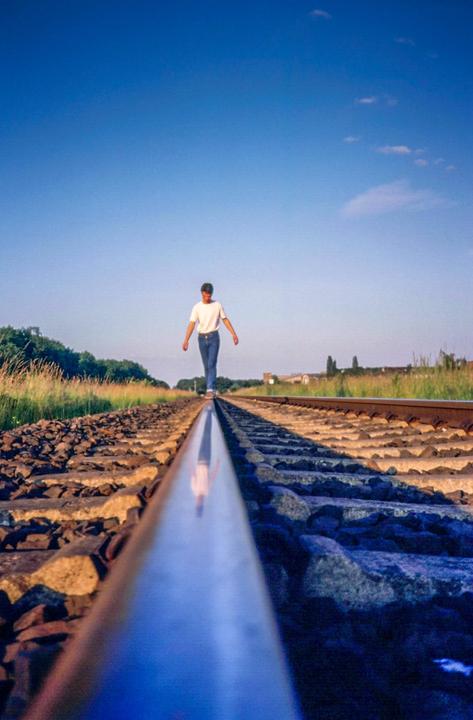 A man walks along a train track with his reflection visible on the rail.