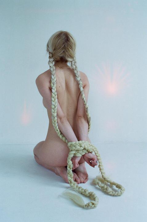 A woman kneels with her hands tied behind her back by her own braided hair.