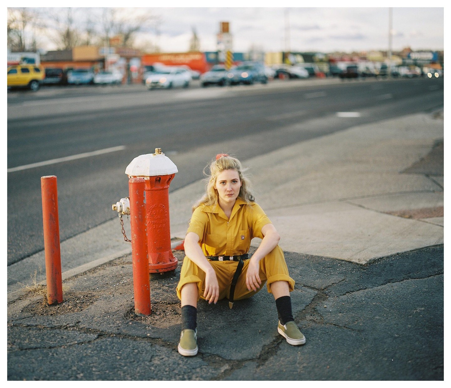 A woman in a yellow jumpsuit sits on the ground next to a red fire hydrant on a street with cars in the background.
