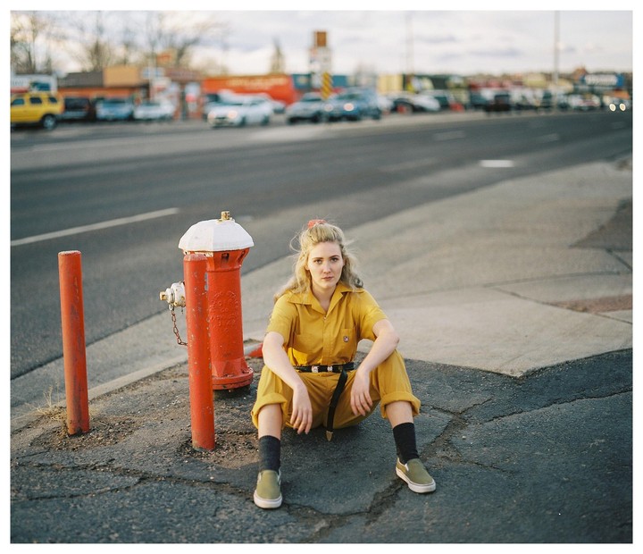 A woman in a yellow jumpsuit sits on the ground next to a red fire hydrant on a street with cars in the background.