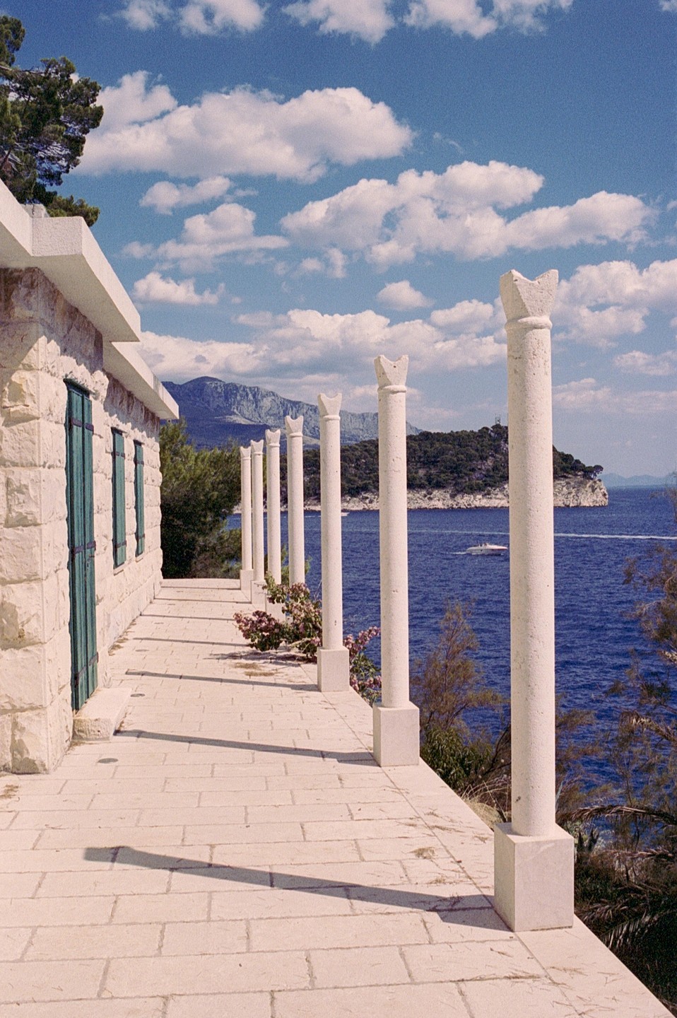 A stone building with green shutters and a row of white pillars overlooks a blue sea with a distant island and mountains under a cloudy sky.