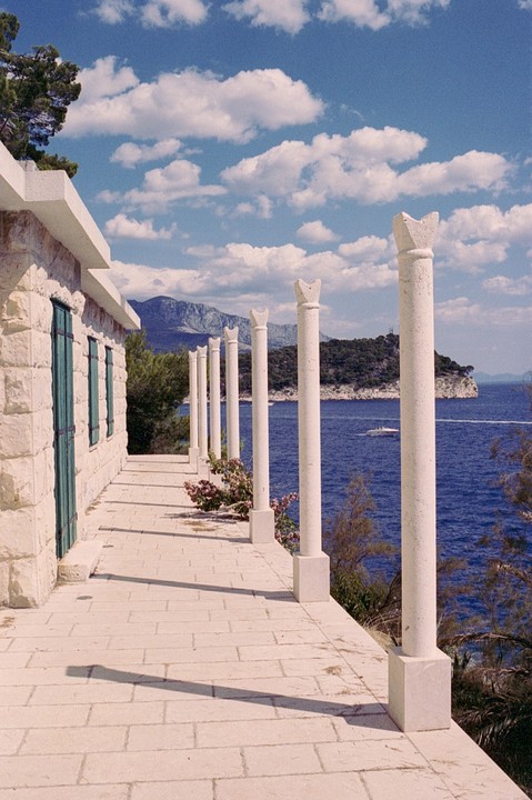 A stone building with green shutters and a row of white pillars overlooks a blue sea with a distant island and mountains under a cloudy sky.