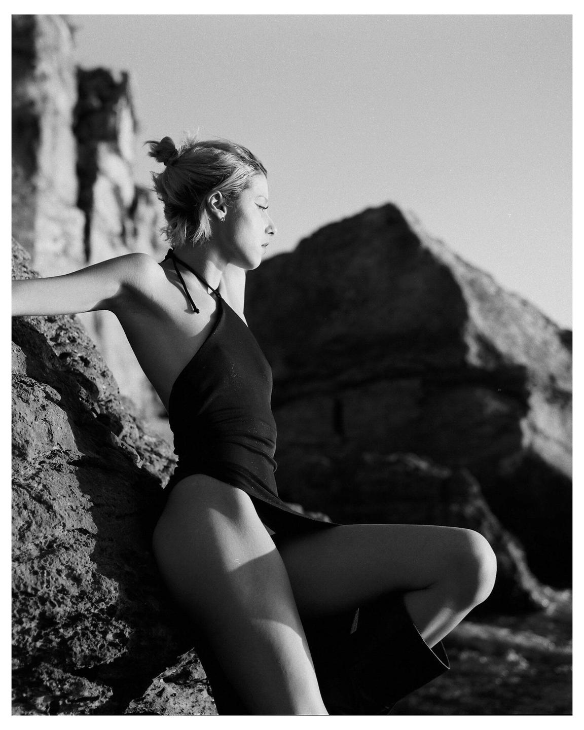 A woman in a dark dress poses on a rocky coast.