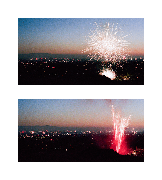 Fireworks explode over a city at dusk, with mountains visible in the distance.