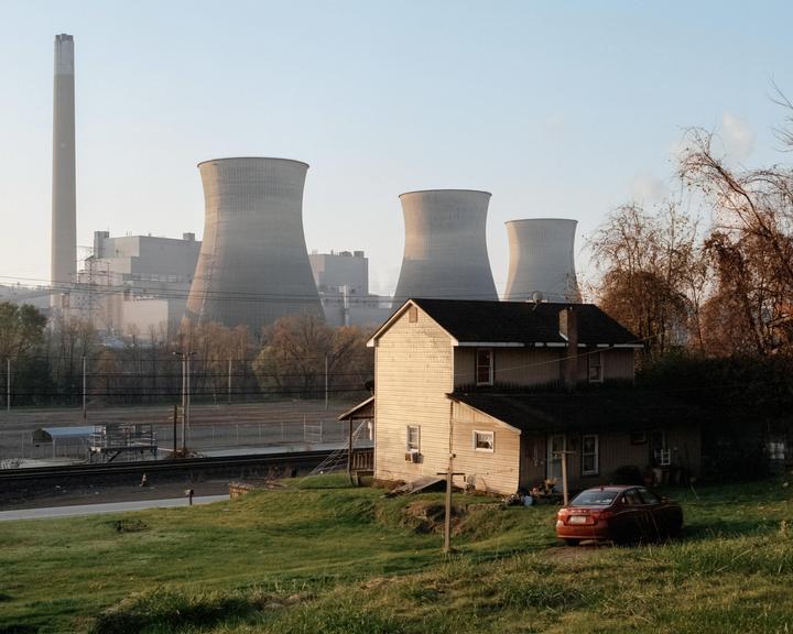 A house and a car are in the foreground, with a power plant and cooling towers in the background.