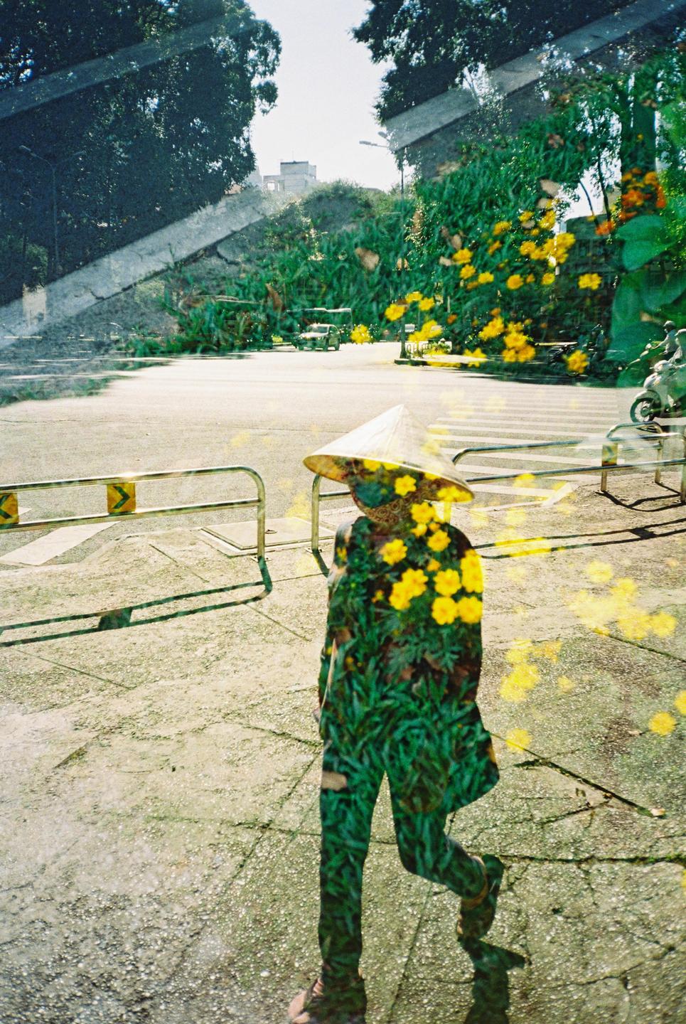 A double exposure shows a person wearing a conical hat walking on a street, overlaid with an image of yellow flowers and green foliage.