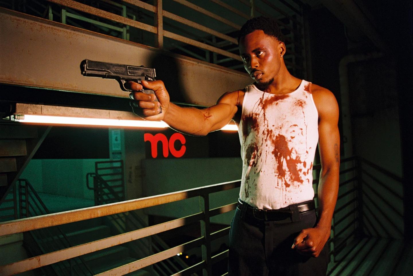 A man in a blood-stained tank top holds a gun in a dimly lit industrial setting.